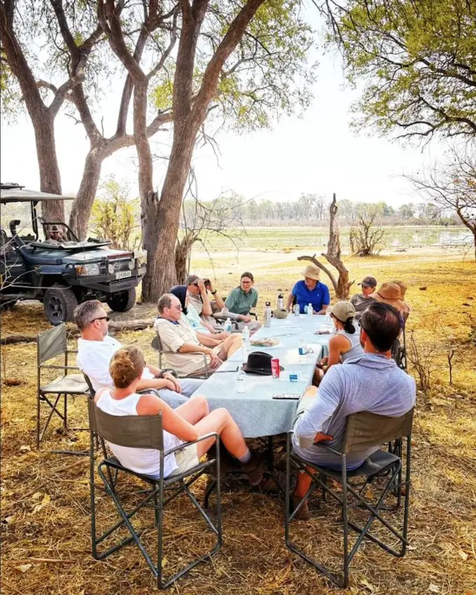 Group dining at outdoor table beneath trees by river with safari vehicle