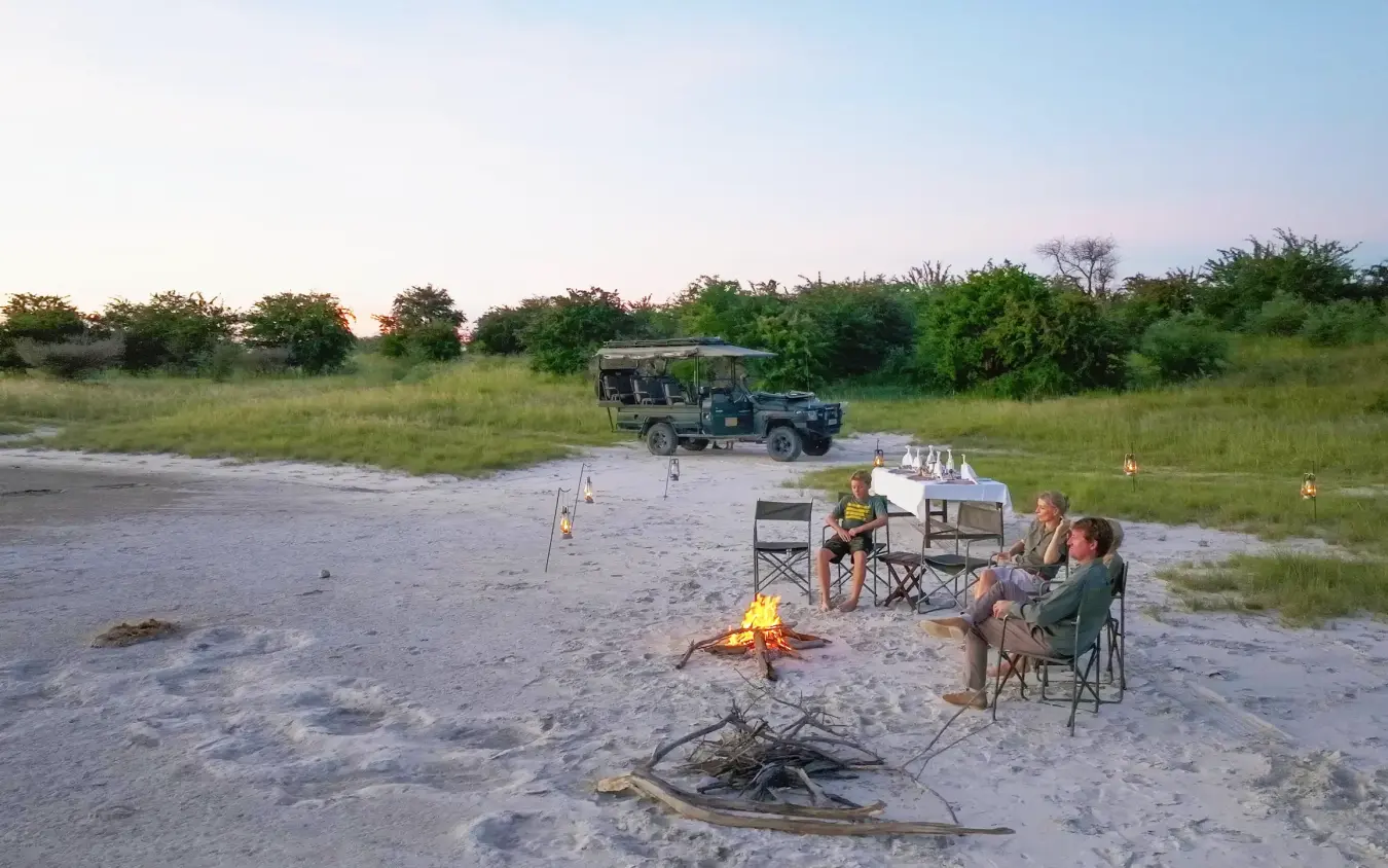 Guests gathered around campfire at safari camp with vehicle parked nearby