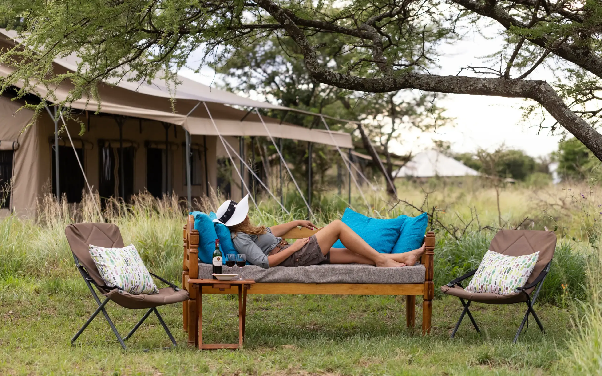 Guest relaxing on outdoor sofa under acacia tree near safari tent, Serengeti Wilderness Camp, Serengeti National Park, Tanzania