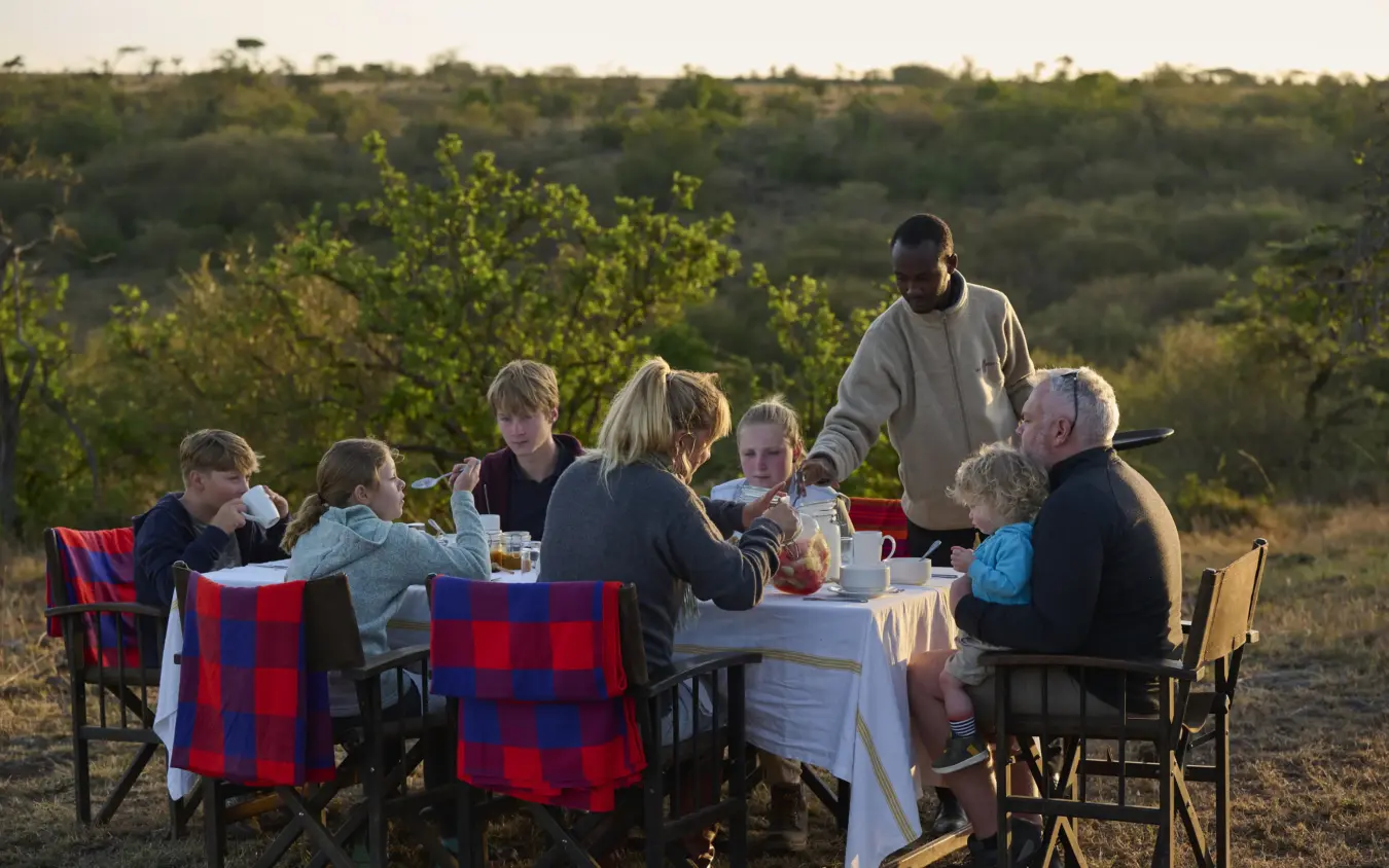 Family dining at outdoor table in savanna landscape at dusk, Basecamp Wilderness, Maasai Mara, Kenya