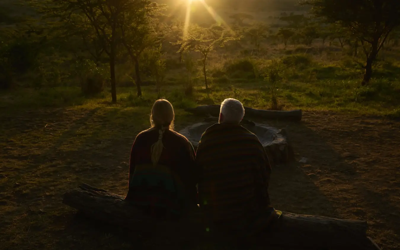 Two people sitting by campfire at dusk in woodland, Basecamp Wilderness, Maasai Mara, Kenya