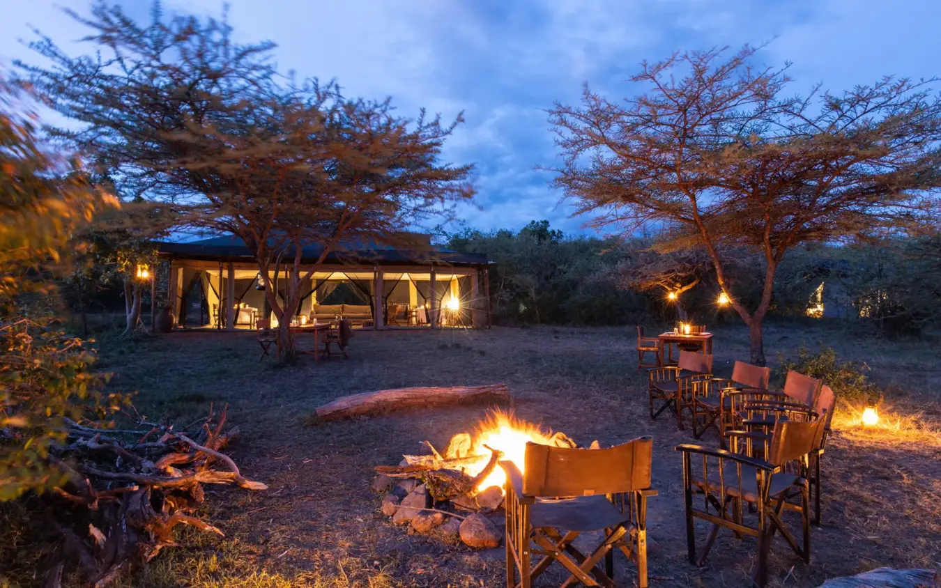 Campfire with chairs and illuminated safari lodge at dusk, Basecamp Wilderness, Maasai Mara, Kenya