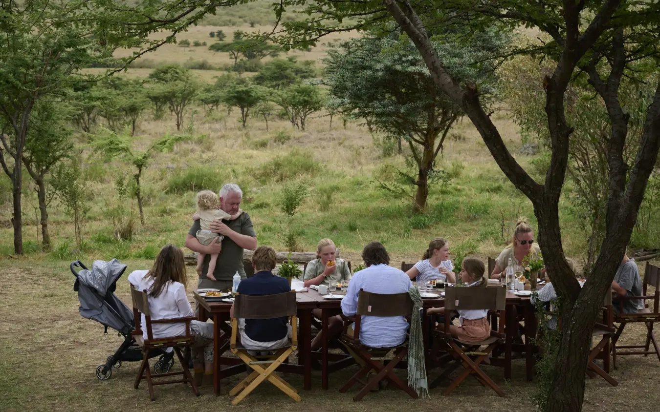 Extended family dining at long table under acacia trees in savanna, Basecamp Wilderness, Maasai Mara, Kenya