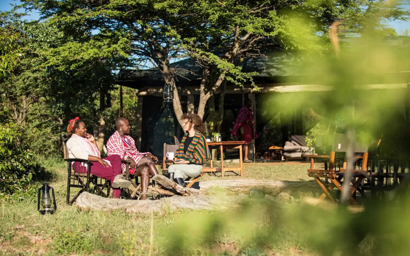 Guests relaxing in camp chairs under acacia tree near lodge buildings, Basecamp Wilderness, Maasai Mara, Kenya