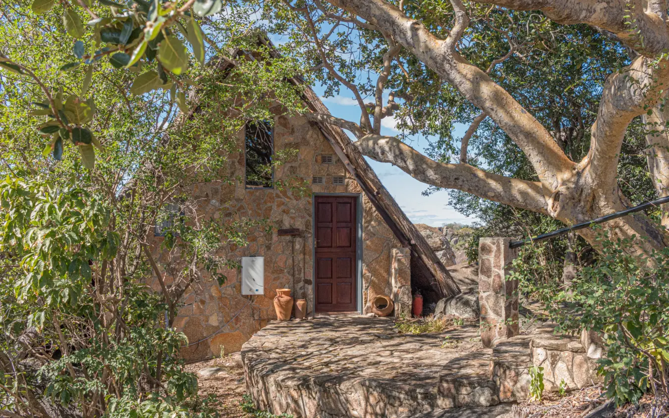 Stone cottage with large tree and wooden door in arid landscape, Big Cave Camp, Matobo, Zimbabwe