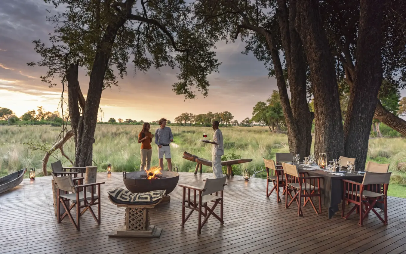 Guests gathered around fire pit on wooden deck beneath ancient trees at dusk, Duba Explorers Camp, The Okavango Delta, Botswana