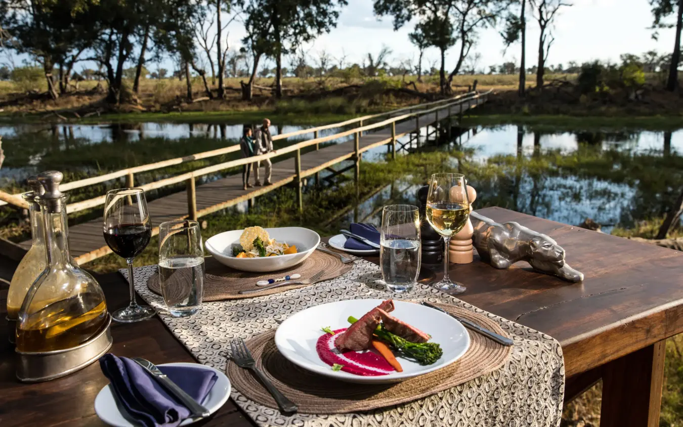 Outdoor dining table with wine and plated meal overlooking wooden bridge and wetland, Duba Explorers Camp, The Okavango Delta, Botswana