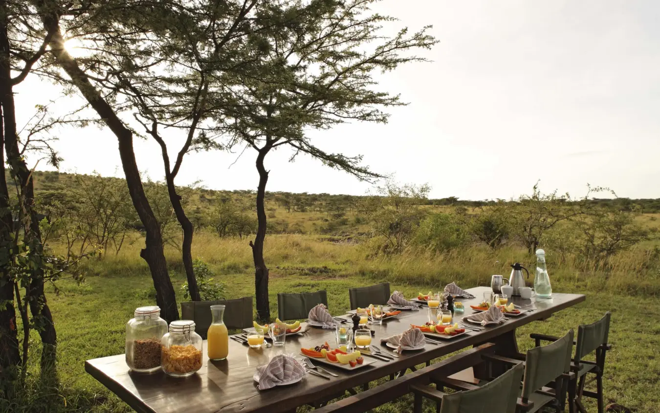 Outdoor dining table set under acacia trees in savanna grassland, Hemingways River Camp Mara, Maasai Mara, Kenya
