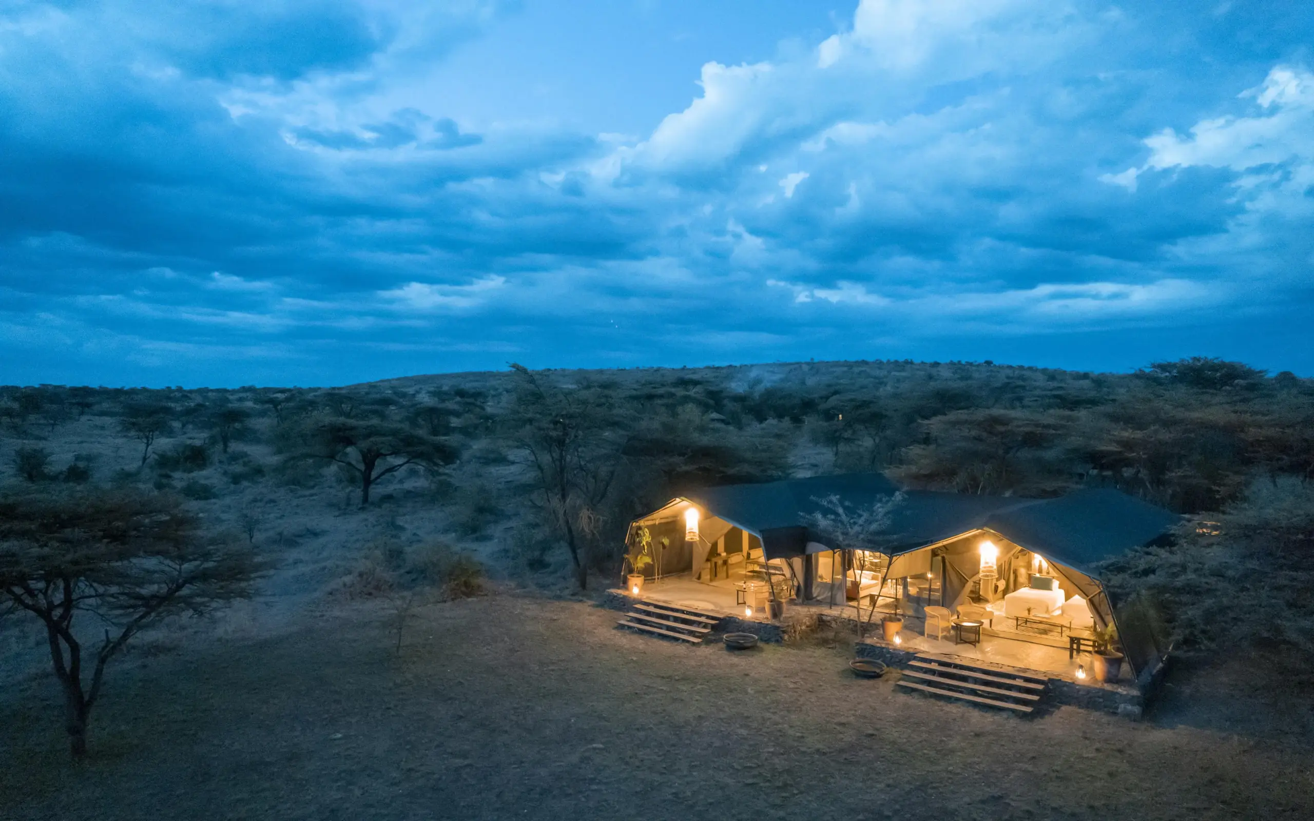 Illuminated safari lodge tents beside waterhole at dusk in arid landscape, Hemingways River Camp Mara, Maasai Mara, Kenya
