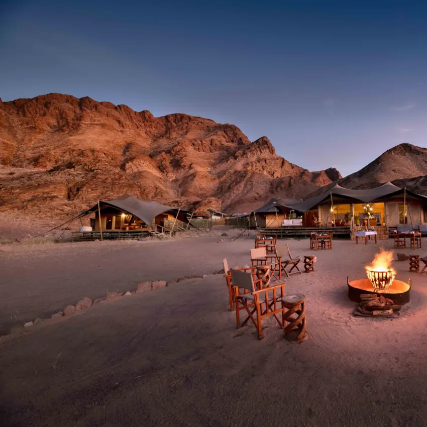 Desert camp with fire pits and canvas tents beneath rocky mountains at dusk, Hoanib Valley Camp, Damaraland, Namibia