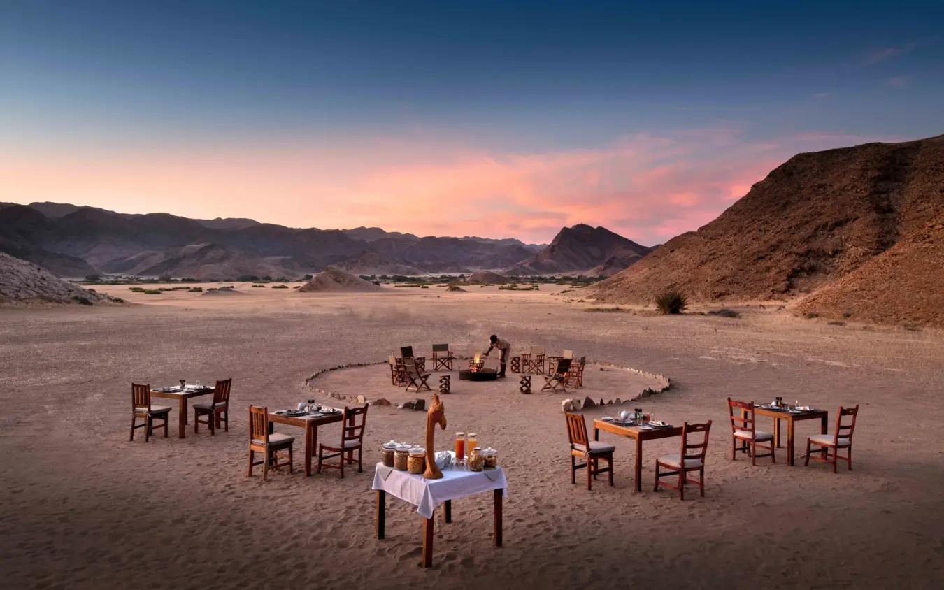 Outdoor dining setup with tables and chairs on desert sand at dusk, Hoanib Valley Camp, Damaraland, Namibia