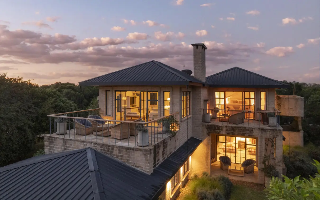Modern stone lodge at dusk with illuminated windows and deck, House In The Wild, Maasai Mara, Kenya