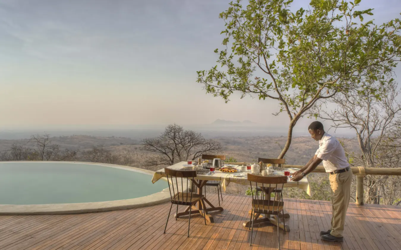 Server setting table for dining on deck overlooking infinity pool and valley landscape, Ikuka Safari Camp, Ruaha National Park, Tanzania