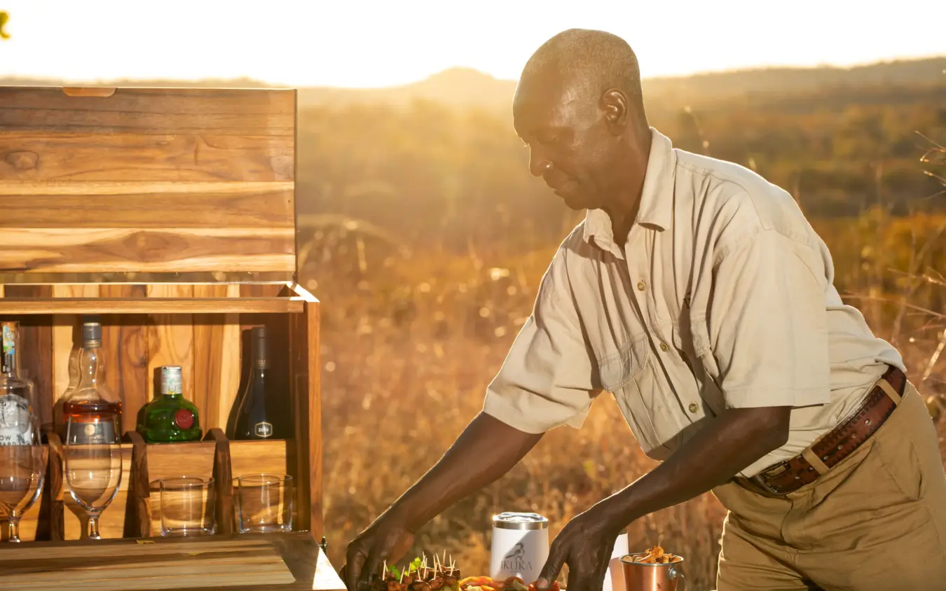 Man arranging drinks at outdoor bar with savanna landscape backdrop, Ikuka Safari Camp, Ruaha National Park, Tanzania