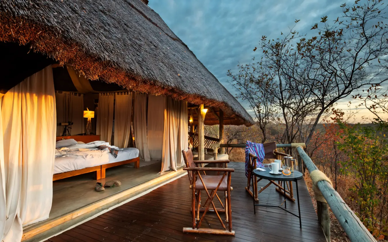 Thatched safari lodge suite with bed and deck overlooking woodland landscape at dusk, Ikuka Safari Camp, Ruaha National Park, Tanzania