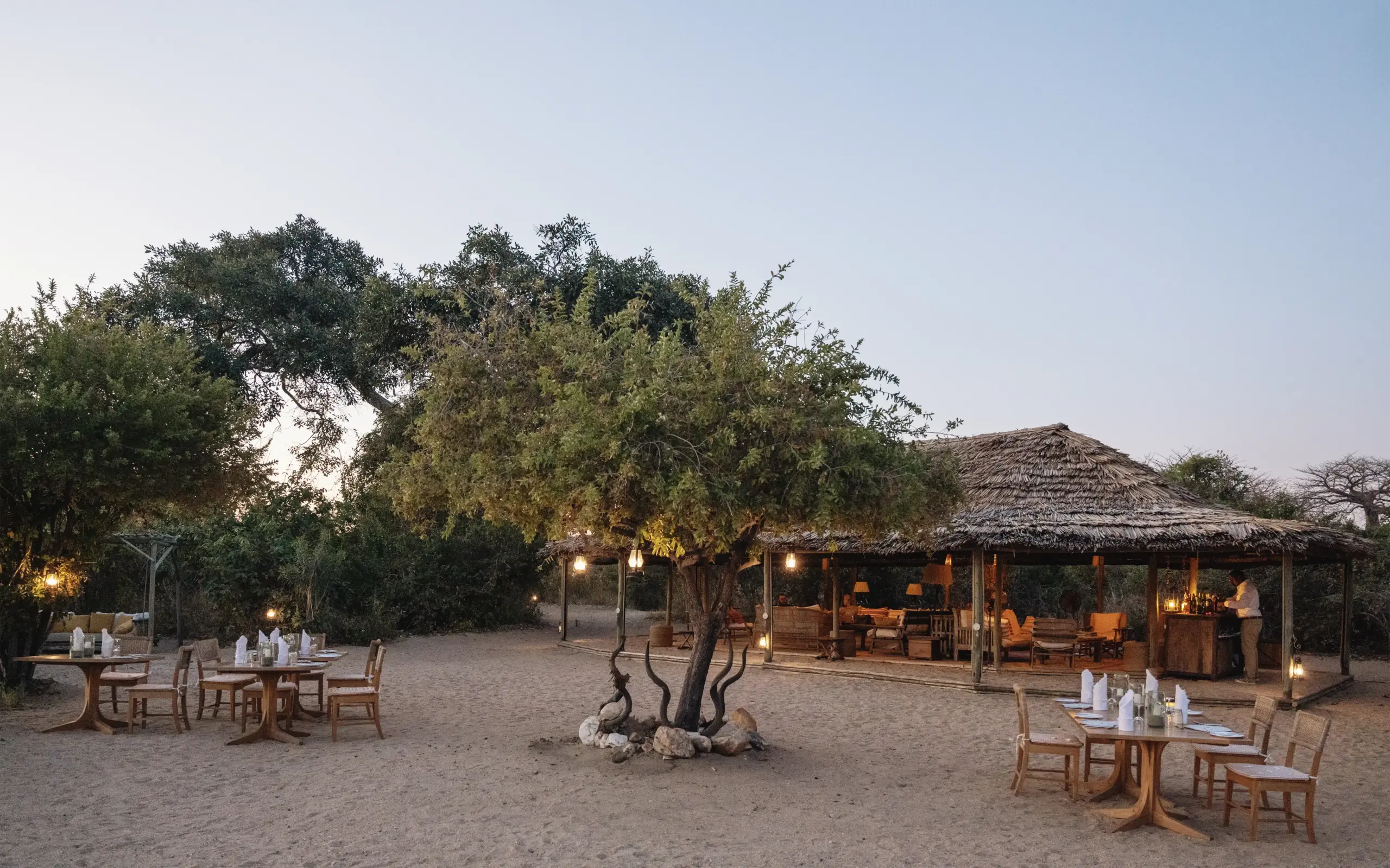 Outdoor dining area with thatched-roof lodge at dusk in sandy setting, Kigelia Ruaha, Ruaha National Park, Tanzania