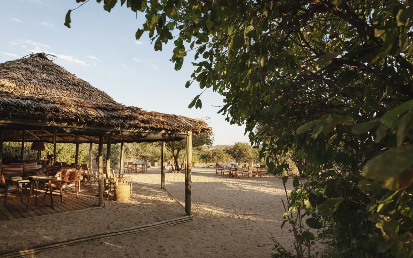 Thatched-roof dining pavilion with wooden furniture on sandy ground at dusk, Kigelia Ruaha, Ruaha National Park, Tanzania