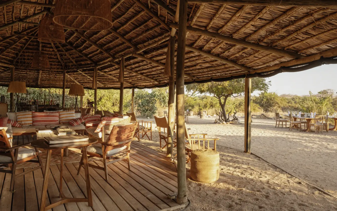 Thatched pavilion with wooden furniture overlooking sandy camping area at dusk, Kigelia Ruaha, Ruaha National Park, Tanzania