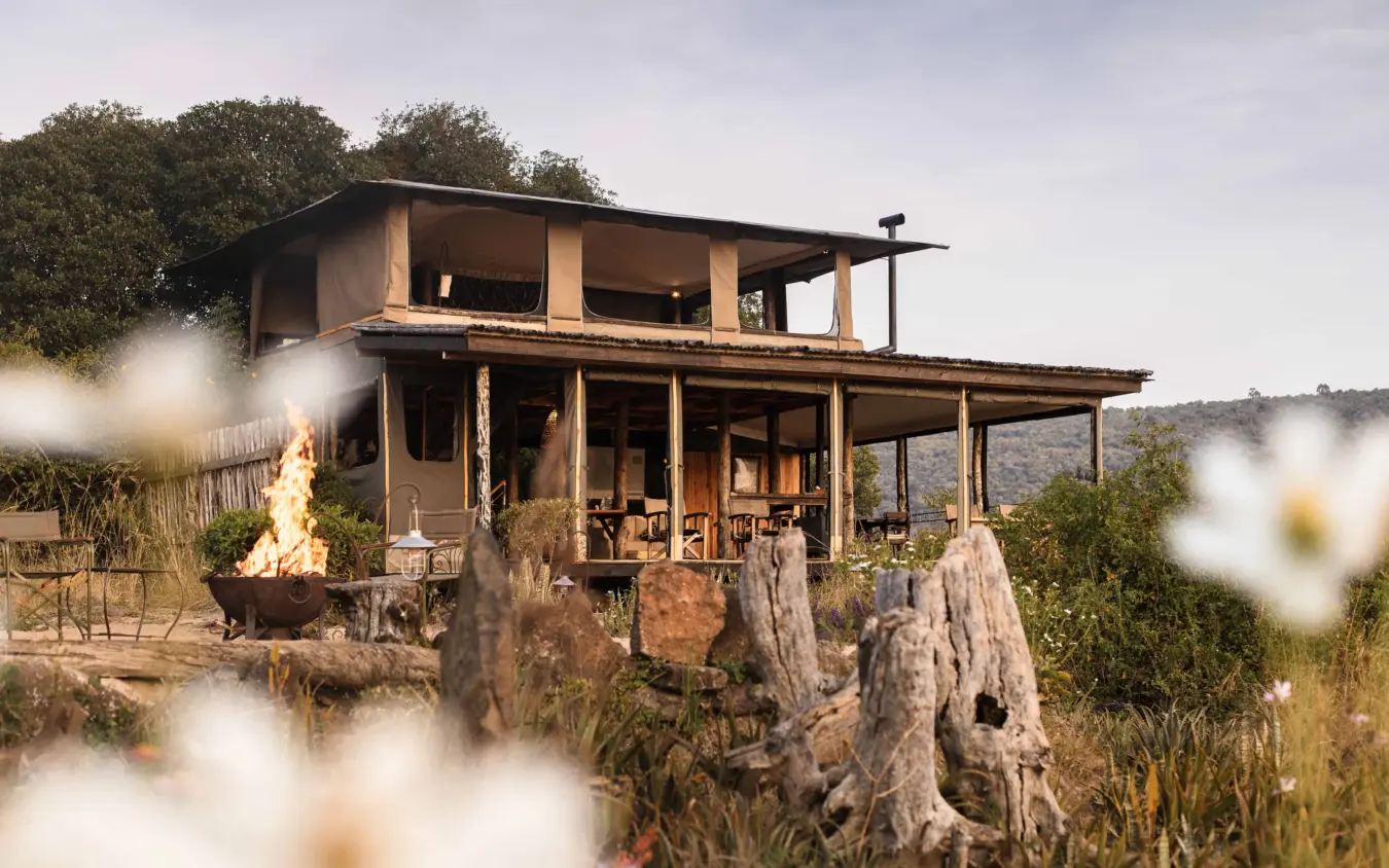 Elevated lodge with fire pit and white flowers in foreground at dusk, Kimya Kimya Camp, Maasai Mara, Kenya