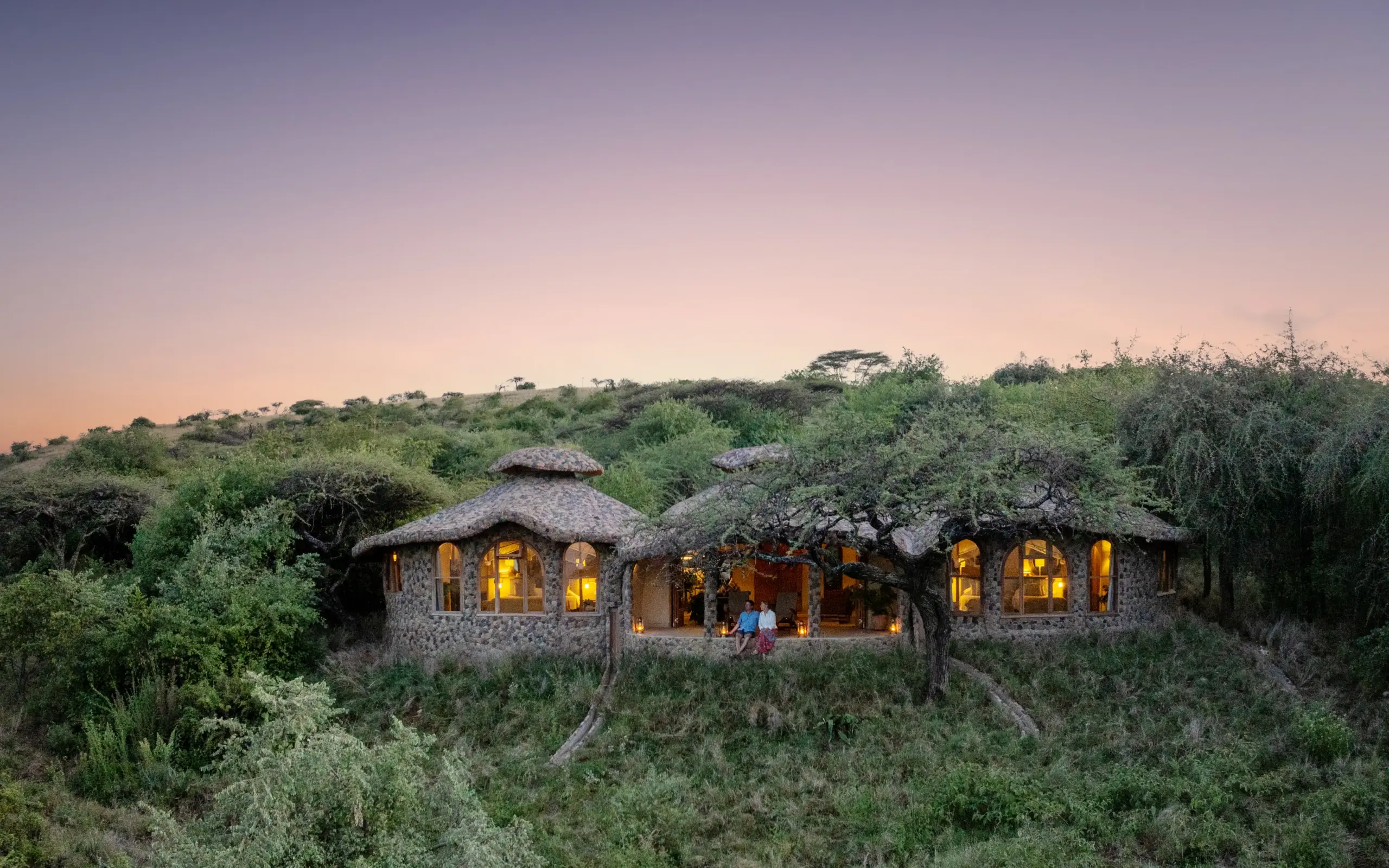 Thatched stone lodge with illuminated windows nestled in scrubland at dusk, Lewa House, Laikipia Highlands, Kenya