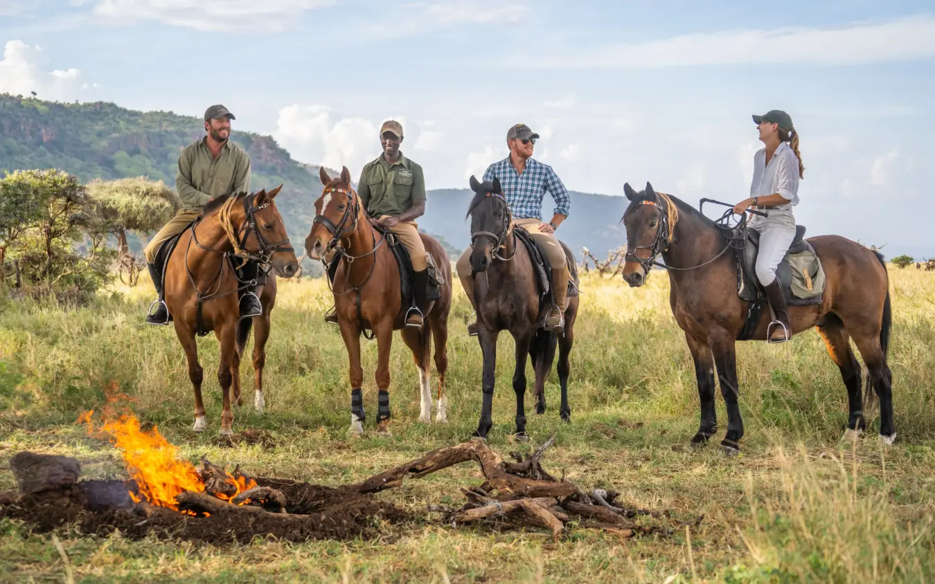 Horseback riders gathered around campfire in grassland with mountain backdrop, Lewa Wilderness, Laikipia Highlands, Kenya