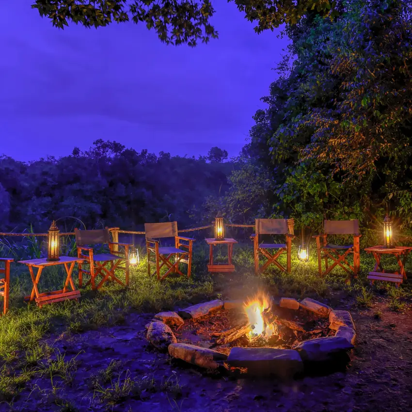 Campfire gathering with canvas chairs and lanterns at dusk in woodland clearing, Naibor Camp, Maasai Mara, Kenya