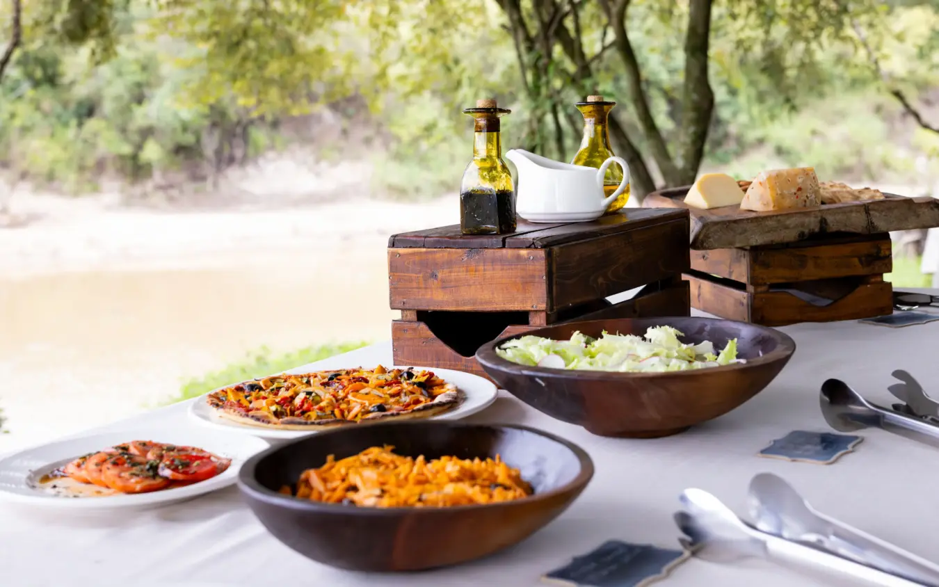 Outdoor dining setup with fresh food and beverages under trees, Naibor Camp, Maasai Mara, Kenya