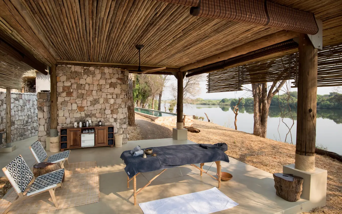 Covered riverside deck with seating and massage table overlooking water, Matetsi River Lodge, Victoria Falls, Zimbabwe