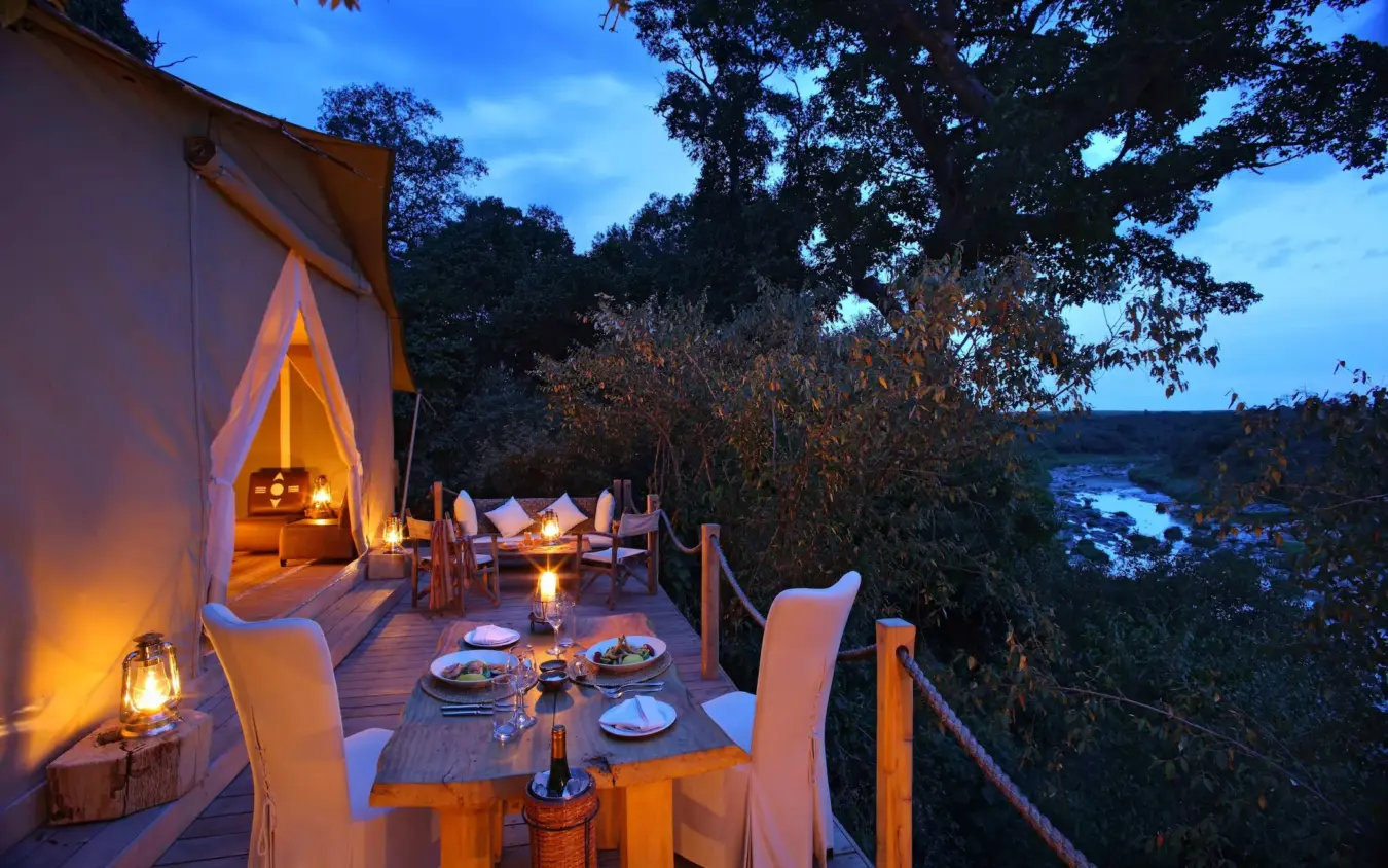 Elevated dining deck overlooking river at dusk with glowing tent, Naibor Camp, Maasai Mara, Kenya