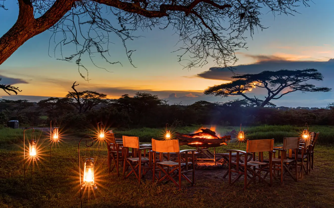 Outdoor dining setup with chairs and campfire under acacia trees at dusk, Ndutu Mobile Tented Camp, Serengeti National Park, Tanzania