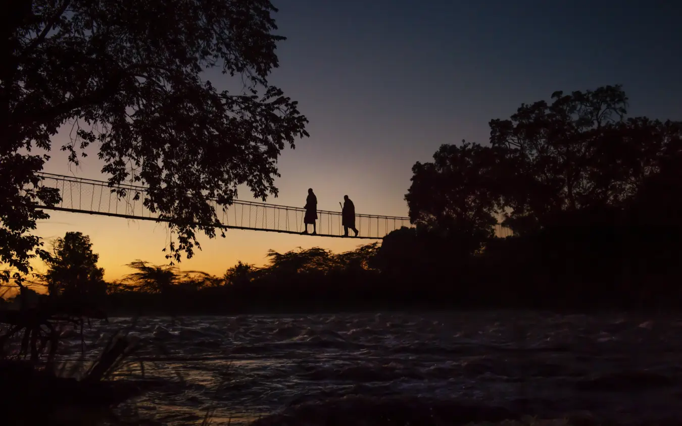 Two figures silhouetted on suspension bridge over river at dusk, Ngare Serian, Maasai Mara, Kenya