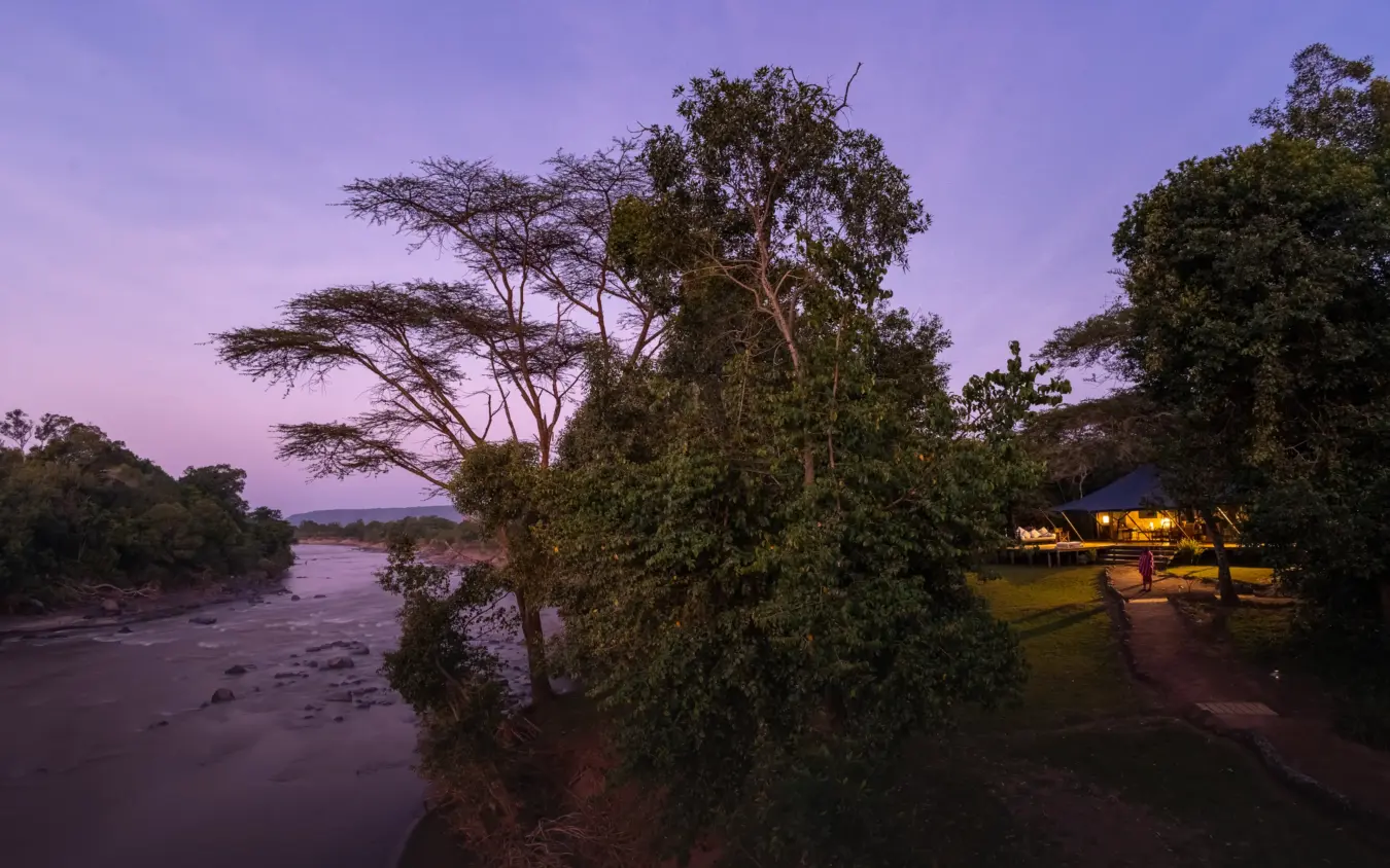 Riverside lodge with illuminated structures at dusk overlooking wide river, Ngare Serian, Maasai Mara, Kenya