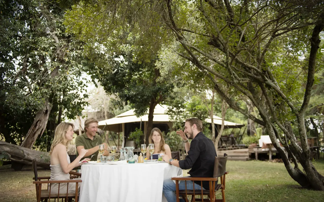 Outdoor dining table under large trees with four guests in natural setting, Ngare Serian, Maasai Mara, Kenya