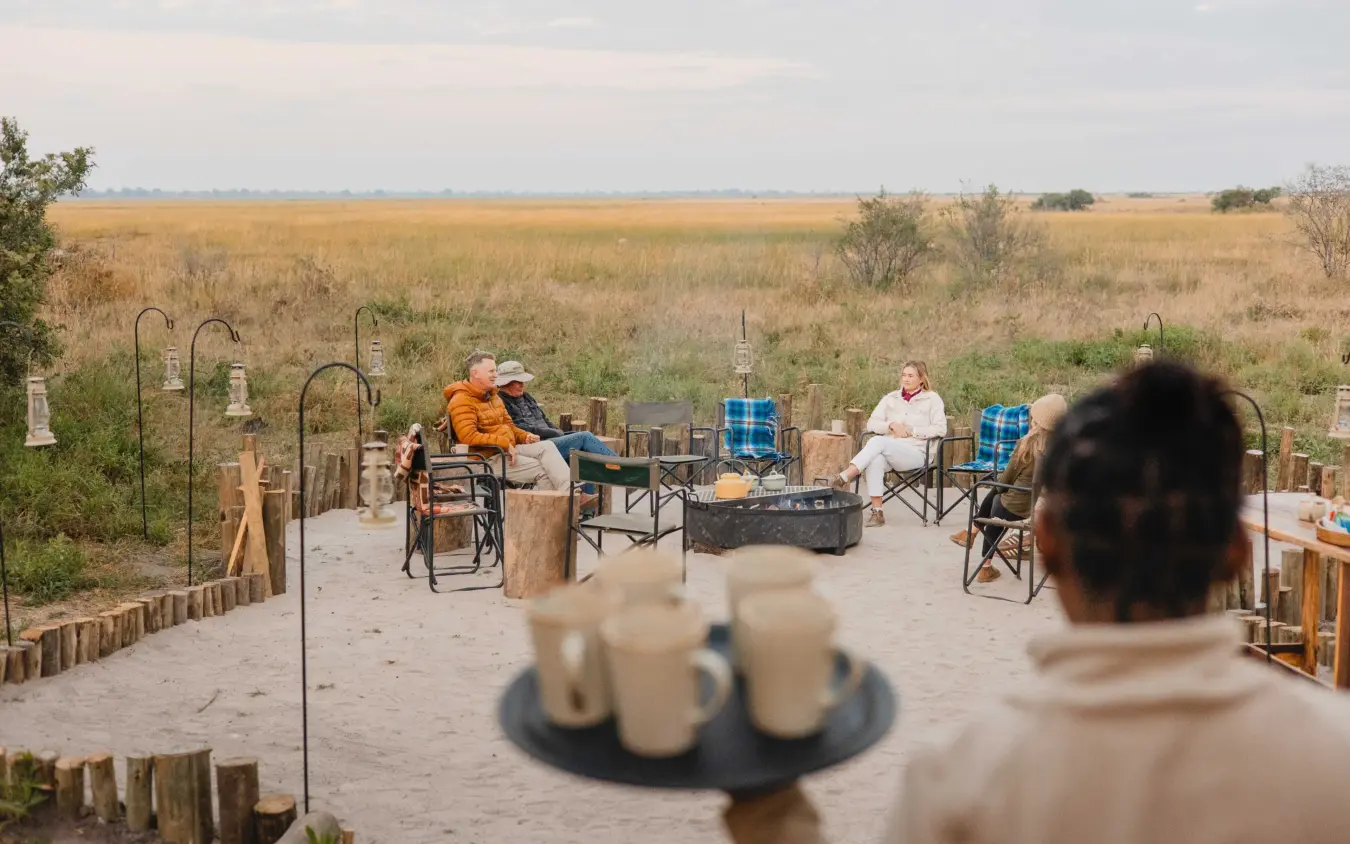 Group gathered around fire pit on sandy terrace overlooking grassland, Nkasa Linyanti, Zambezi Region, Namibia