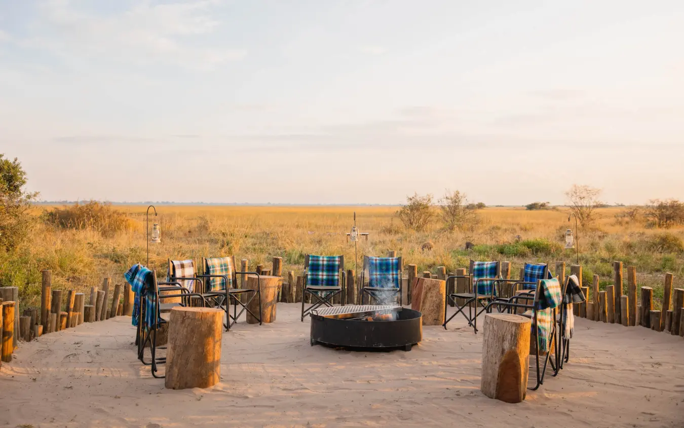 Outdoor lounge area with campfire and chairs overlooking savanna grassland at dusk, Nkasa Linyanti, Zambezi Region, Namibia