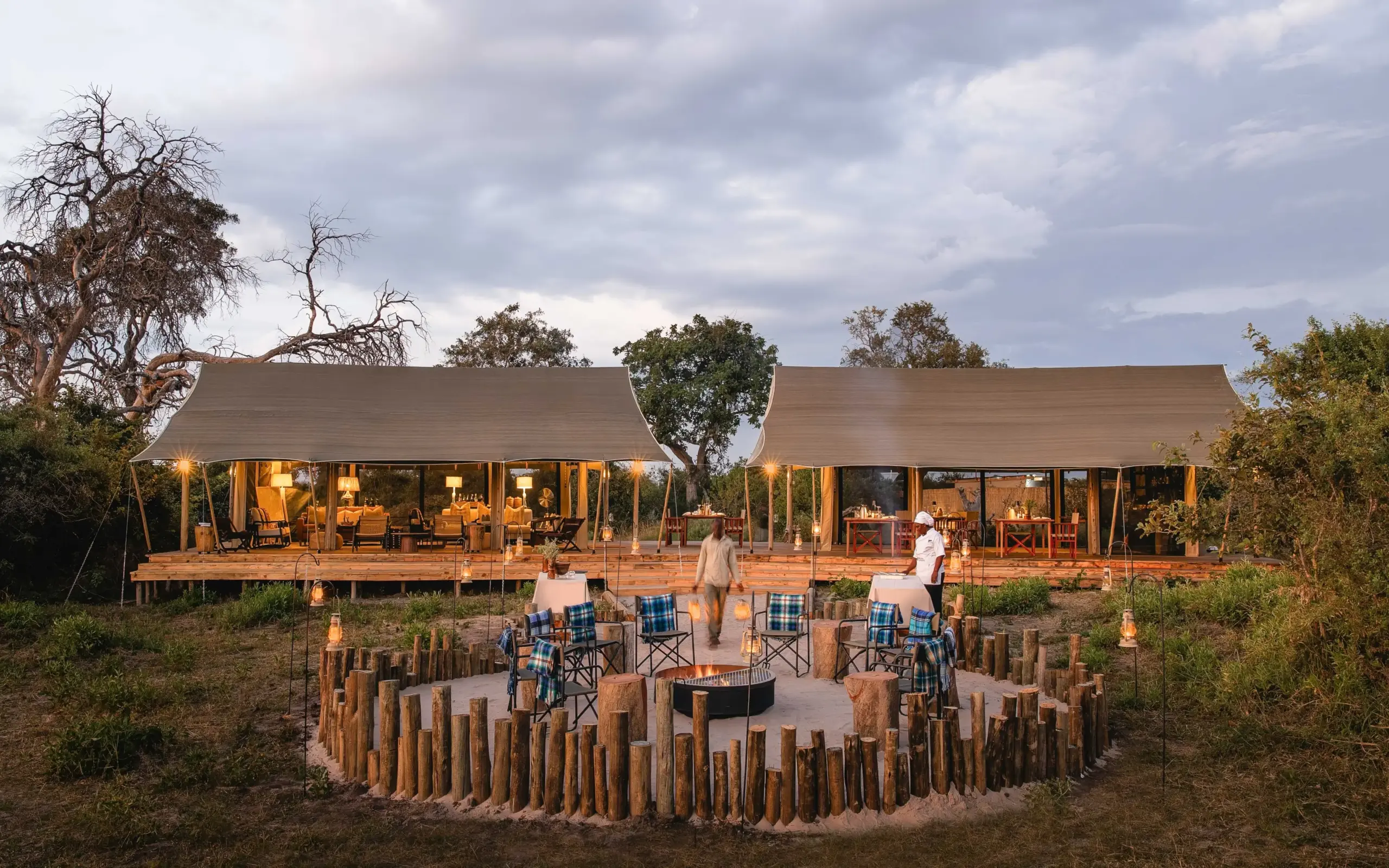 Luxury safari lodge with illuminated interior and outdoor firepit seating at dusk, Nkasa Linyanti, Zambezi Region, Namibia