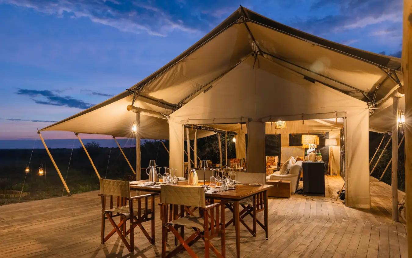 Illuminated canvas safari tent with dining area on wooden deck at dusk, Nkasa Linyanti, Zambezi Region, Namibia