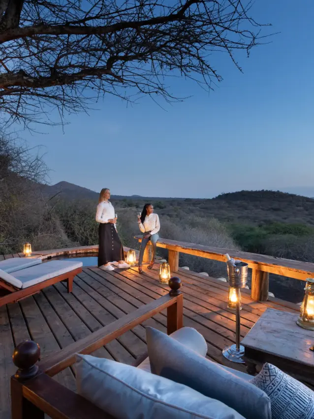 Guests on wooden deck at sunset with savanna landscape and lantern lighting, Ol Donyo Lodge, Amboseli and Chyulu Hills, Kenya