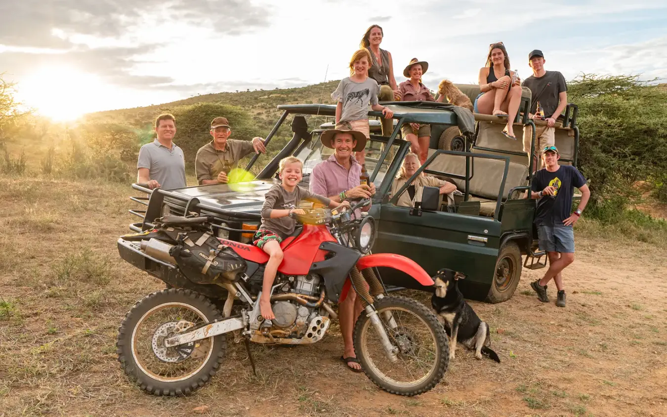 Safari vehicle and motorcycle with group posing at sunset in savanna landscape, Ol Malo House, Laikipia Highlands, Kenya