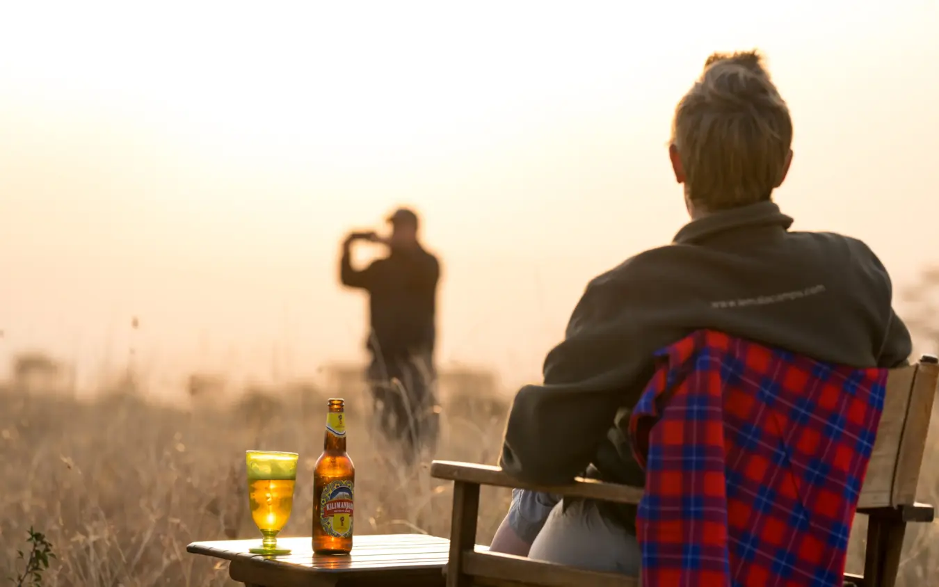 Woman with beer watching friend photograph sunset over dry grassland, Osonjoi Lodge, Ngorongoro Crater, Tanzania