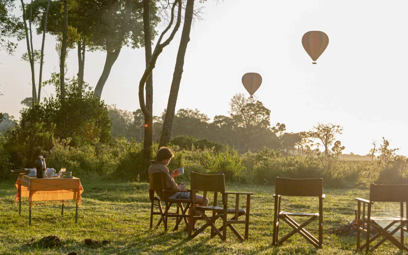 Outdoor breakfast setup with hot air balloons rising above savanna landscape, Roca River Camp, Maasai Mara, Kenya