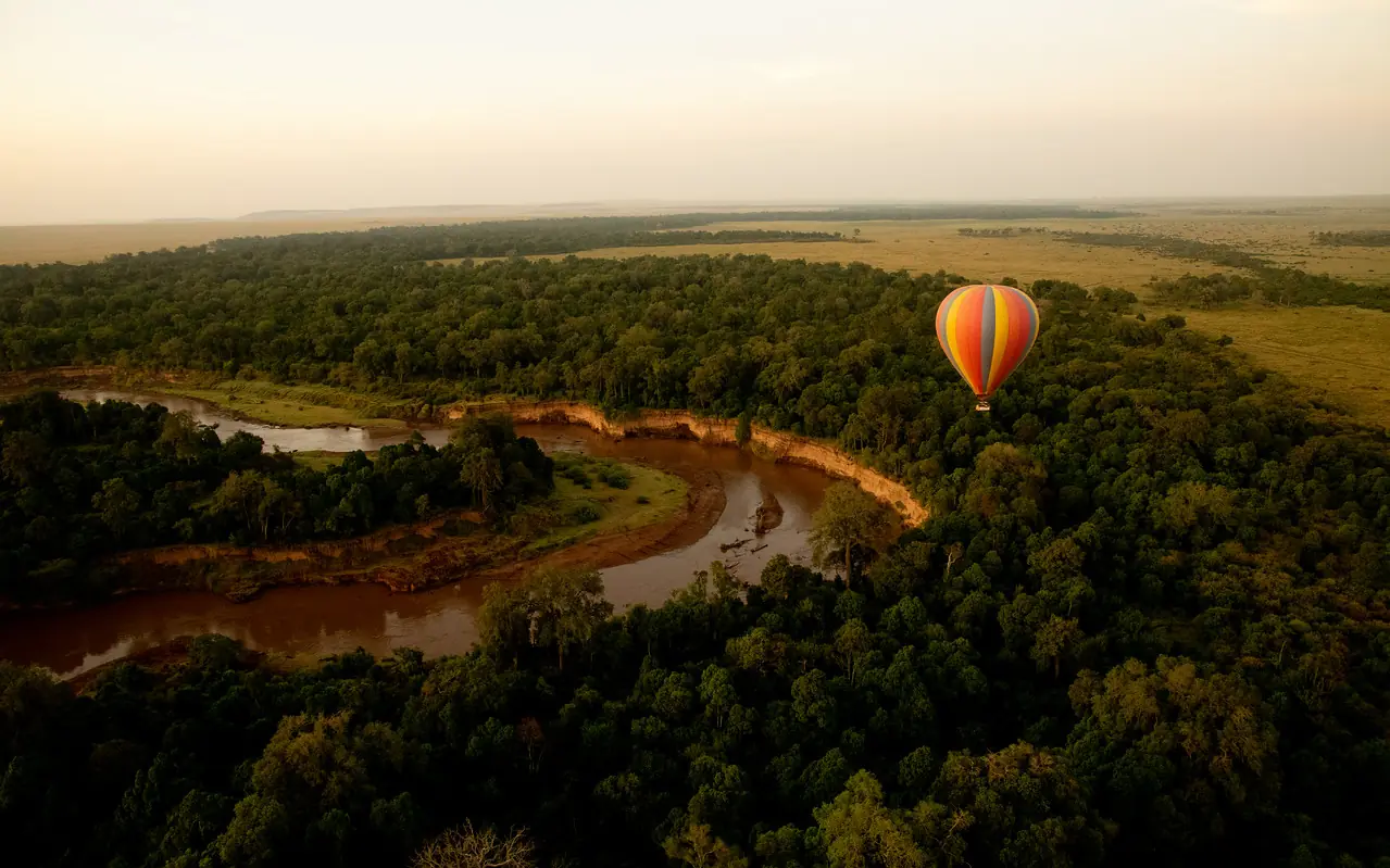 Hot air balloon floating above meandering river through dense forest, Roca River Camp, Maasai Mara, Kenya