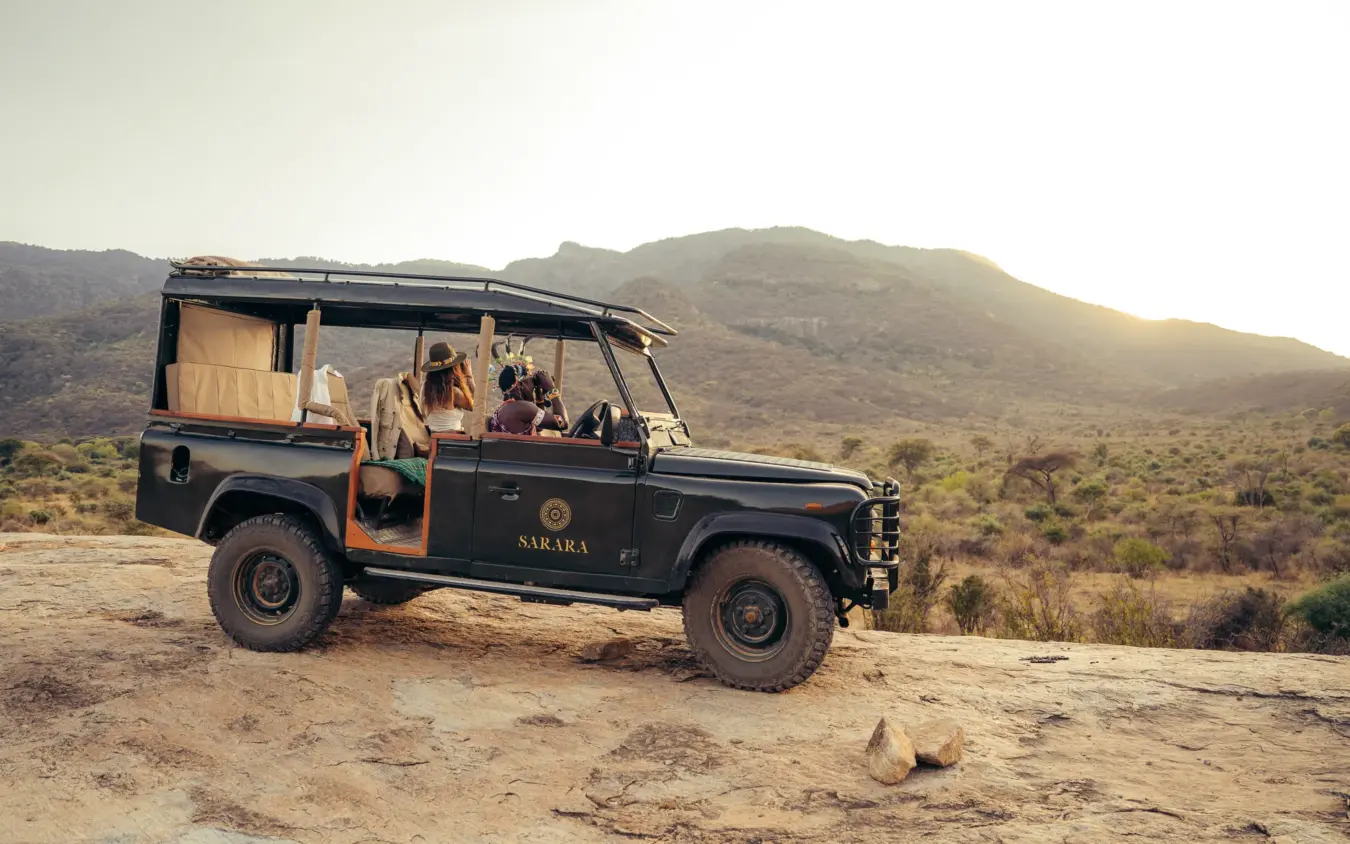 Open-sided safari vehicle with guests on rocky terrain with mountains at sunset, Sarara Camp, Northern Kenya, Kenya