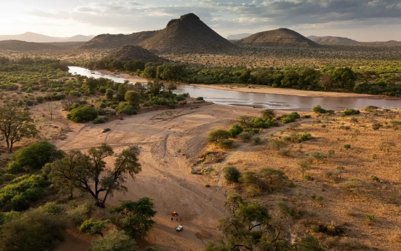 Safari vehicle crossing dry riverbed beneath volcanic peaks, Sarara Wilderness, Northern Kenya, Kenya