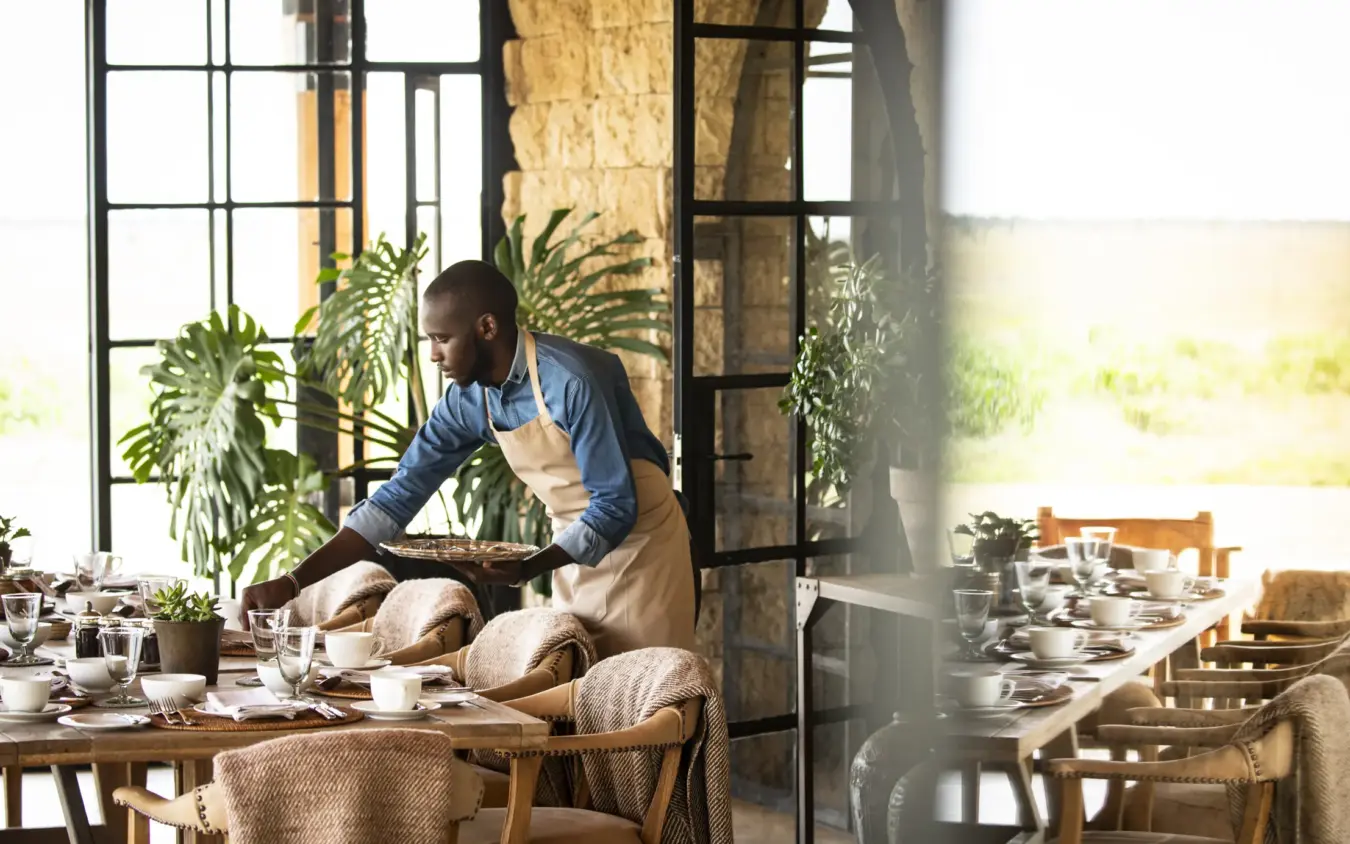 Man arranging fresh bread on dining table with potted plants and windows, Segera, Laikipia Highlands, Kenya