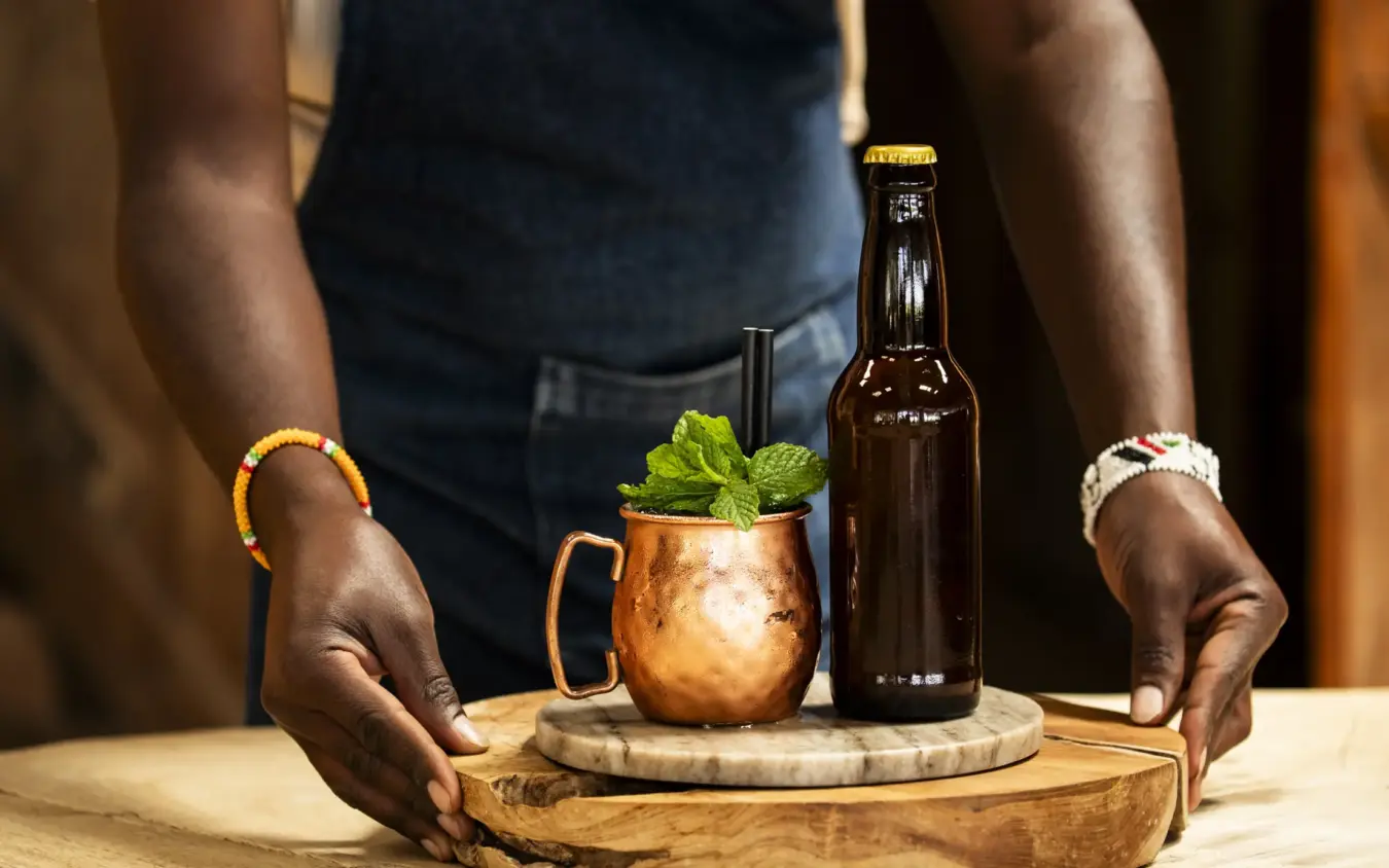 Man presenting Moscow Mule cocktail with beer bottle on wooden table, Segera, Laikipia Highlands, Kenya