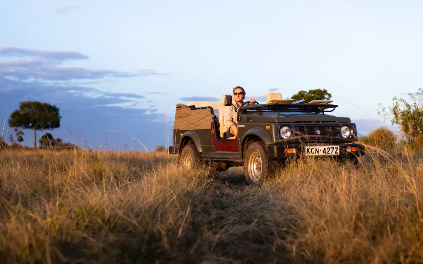 Safari vehicle with guide parked in golden grassland at dusk, Serian “The Original”, Maasai Mara, Kenya
