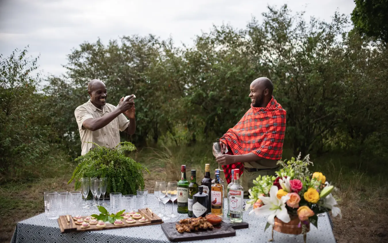 Two men enjoying outdoor picnic with drinks and flowers in natural landscape, Seringet Mara Camp, Maasai Mara, Kenya