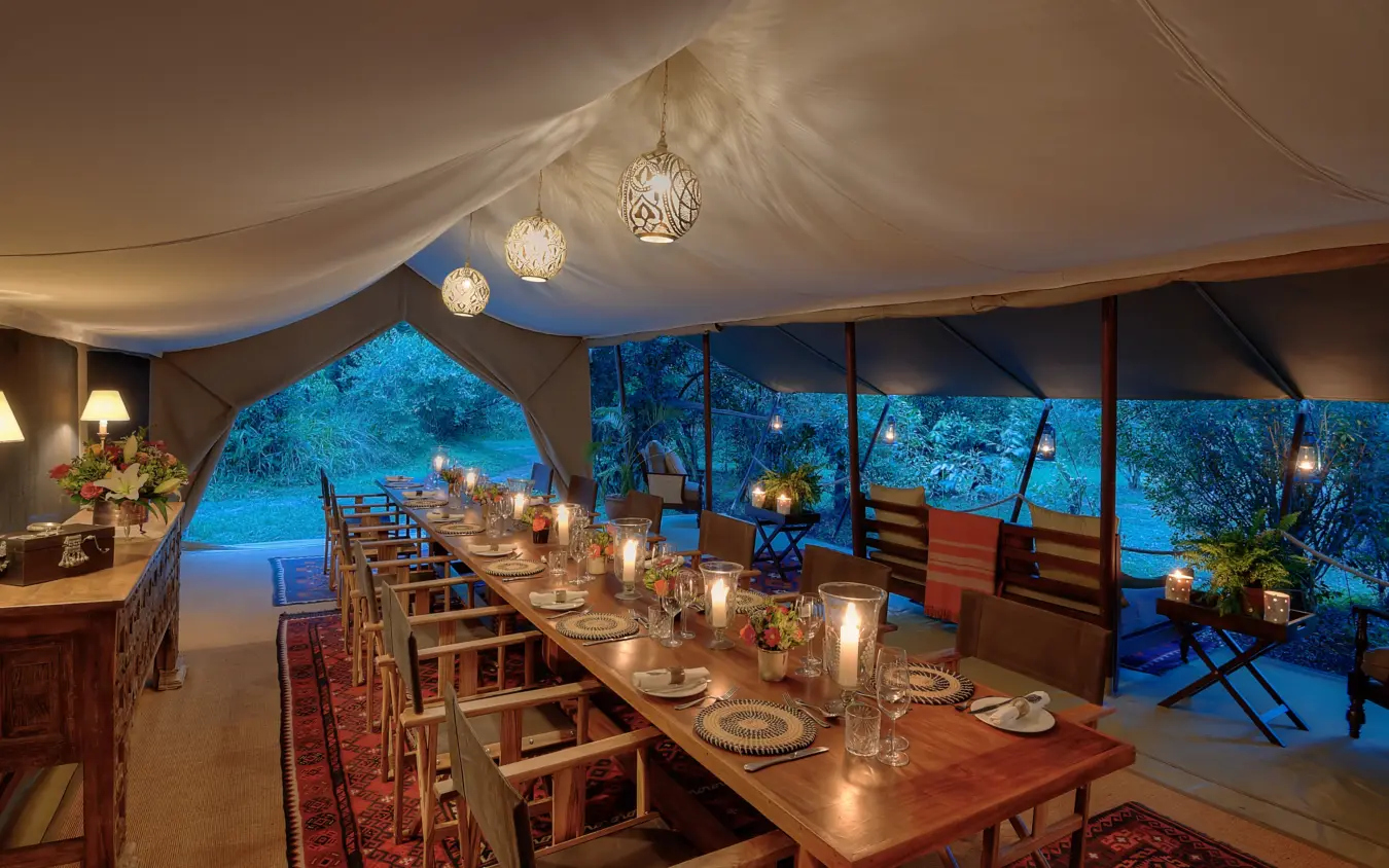 Candlelit dining tent with floor-to-ceiling windows overlooking illuminated landscape at dusk, Seringet Mara Camp, Maasai Mara, Kenya