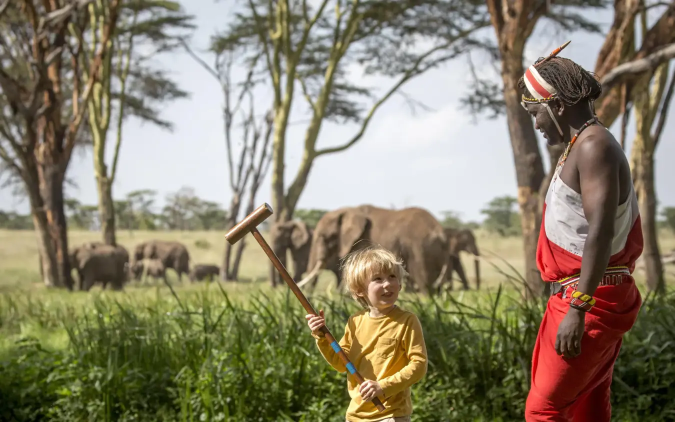 Child playing croquet with Maasai guide, elephants in background, Sirikoi, Laikipia Highlands, Kenya